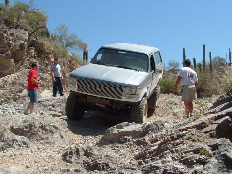 1990 Ford Bronco - Silver 1990 Ford Bronco tackling a rough, rocky trail in a desert landscape.