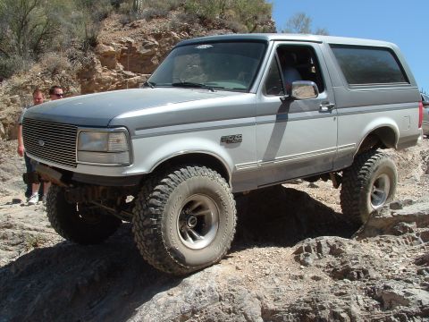 1990 Ford Bronco - A lifted Ford Bronco navigates a rocky trail under a clear blue sky.