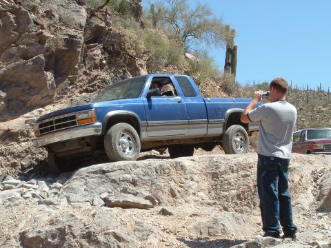 1990 Ford Bronco - A blue Dodge pickup truck navigates a rocky off-road trail, with a man photographing the scene.