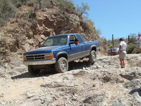 1990 Ford Bronco - A blue Dodge Dakota navigates a rocky off-road trail, with another vehicle and people in the background.