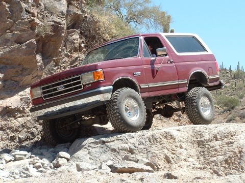 1990 Ford Bronco - A maroon Ford Bronco navigates a rocky trail in a desert landscape under a clear blue sky.