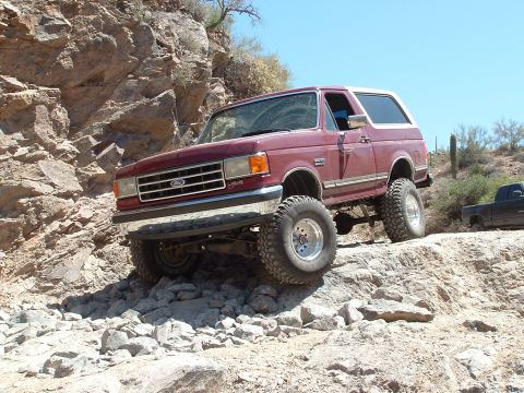 1990 Ford Bronco - A maroon Ford Bronco navigates a rocky off-road trail under a clear blue sky.