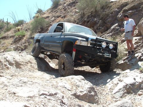 1990 Ford Bronco - A man watches a modified pickup truck crawl over rocks on a desert trail.