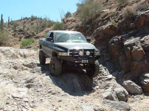 1990 Ford Bronco - Modified pickup truck driving on a rough, rocky desert trail with cacti and scrub brush.