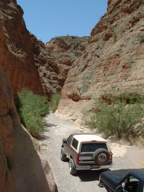 1990 Ford Bronco - A brown Ford Bronco navigates a narrow, rocky canyon on a sunny day, with another vehicle partially visible in the foreground.
