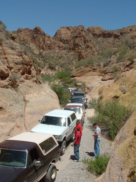 1990 Ford Bronco - A line of SUVs, including a white Ford Bronco, navigates a narrow, rocky canyon during an off-road excursion.