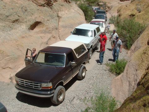 1990 Ford Bronco - A line of SUVs, including a brown 1990 Ford Bronco, navigates a narrow dirt road through a rocky canyon.