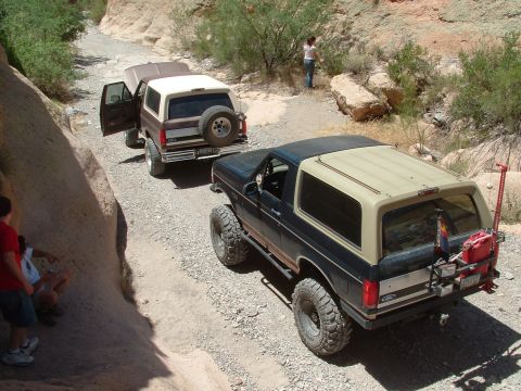 1990 Ford Bronco - Two Ford Broncos navigate a rocky canyon trail, kicking up dust as they go.