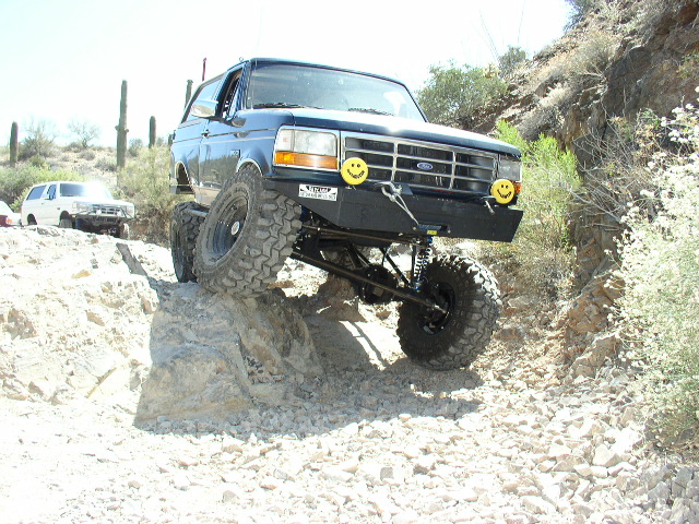 Black lifted Ford Bronco climbing rock ledge on desert trail, front tire lifted