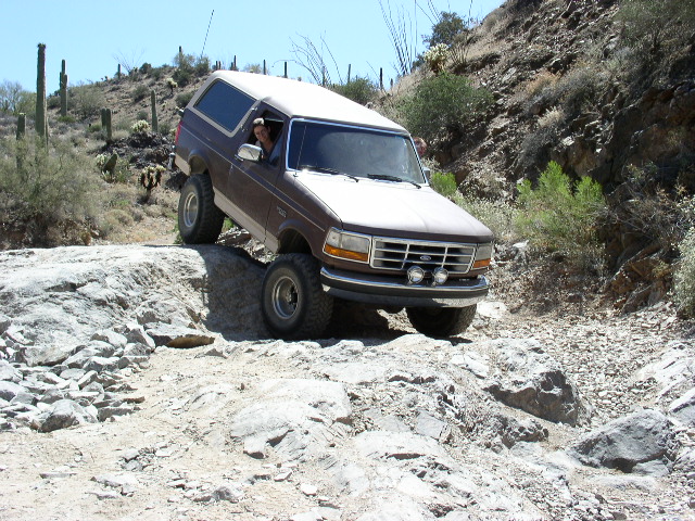 Brown and tan Ford Bronco with white top on rocky desert trail among saguaro cactus