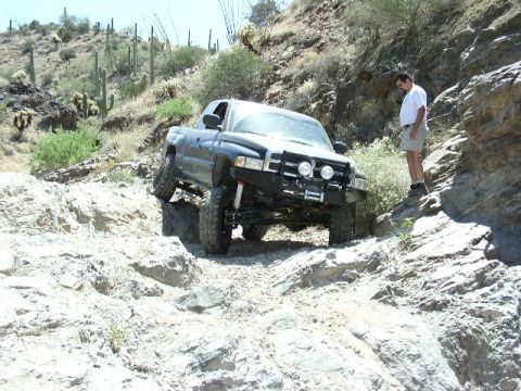 1993 Ford Bronco - A black pickup truck tackles a challenging rocky terrain in a desert environment, with a man observing from the side.
