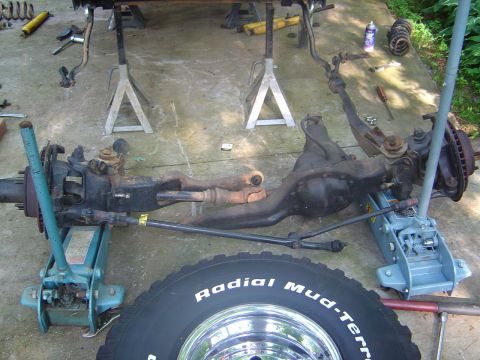 1986 Ford Bronco - A removed front axle assembly (Dana solid axle with differential, tie rod, drag link and knuckles) sits on the concrete, supported by two f…