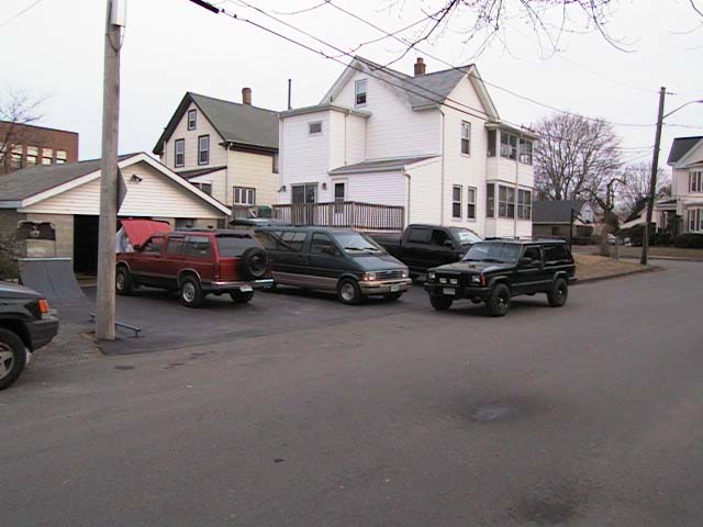 Black Jeep Cherokee XJ parked with a maroon pickup and minivan by a white house