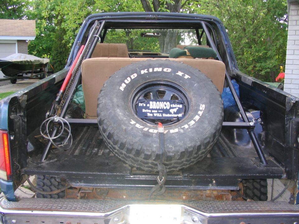 Spare tire mounted in the bed of a dark blue SUV, with a "Bronco thing" license plate and "Mud King XT" tire. Roll cage and jack visible