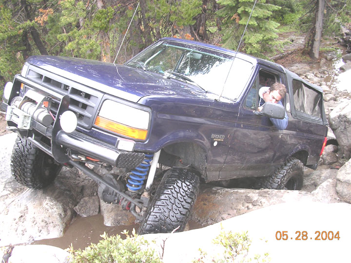 Blue Ford Bronco flexed over rocks on a wooded trail, driver inside, dated photo