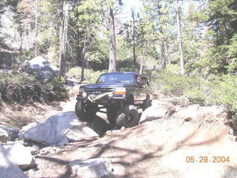 1994 Ford Bronco - A 1994 Ford Bronco navigates a rocky off-road trail surrounded by trees.
