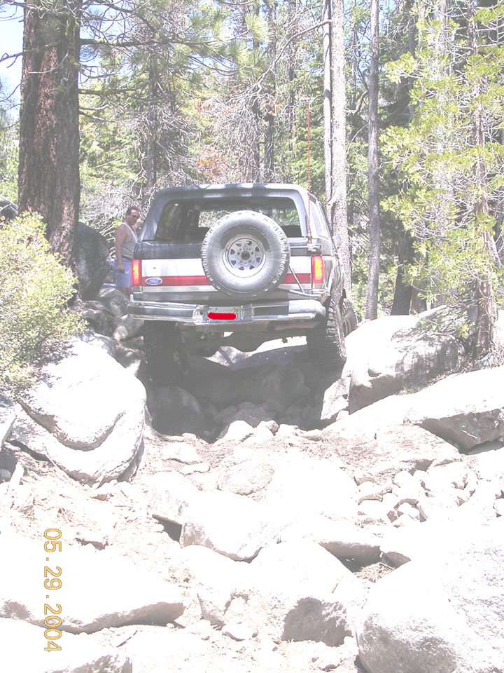 Silver Ford Bronco rear view crawling boulders, tailgate spare and whip antenna, forest trail