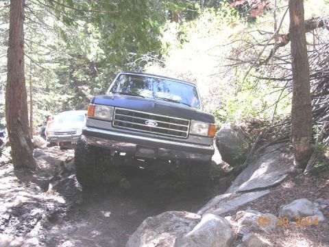 1994 Ford Bronco - Ford Bronco tackling a challenging off-road path with trees and rocks surrounding it.