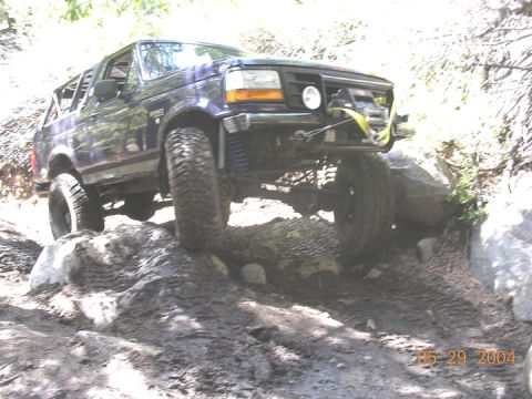 1994 Ford Bronco - A dark blue 1994 Ford Bronco tackles a challenging off-road course with large rocks and mud.