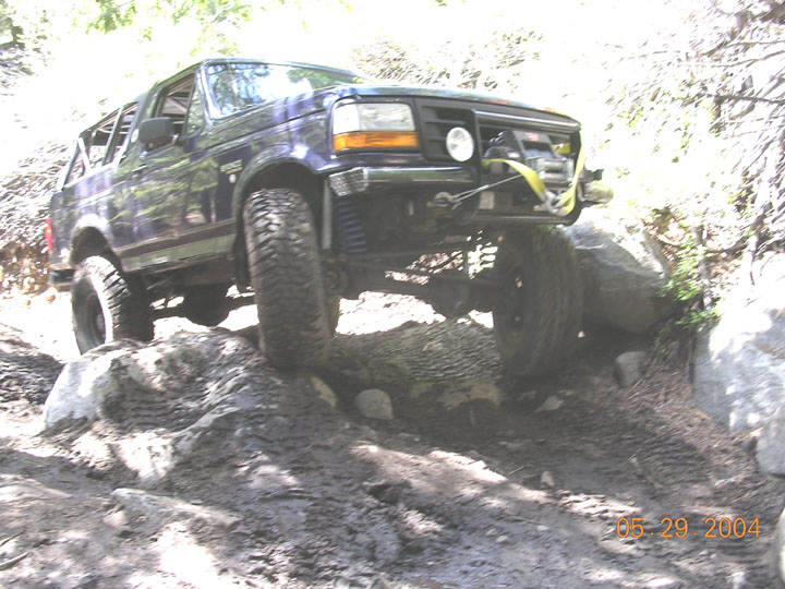 Purple lifted Ford Bronco climbing rocks on a wooded trail with winch bumper