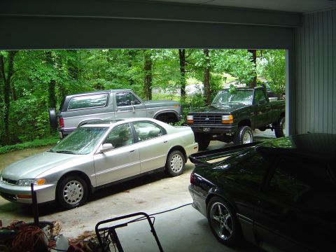 1986 Ford Bronco - View from inside a garage looking out at a wooded driveway, with a silver first-gen full-size Ford Bronco parked at left rear and a dark gr…