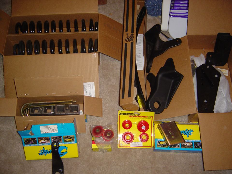 Suspension lift kit parts: black brackets, U-bolts, red poly bushings, and Skyjacker boxes on carpet.