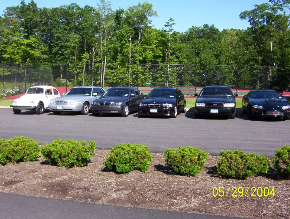 Row of six cars parked outdoors, including a black Ford Crown Victoria, BMWs, and a VW Beetle.