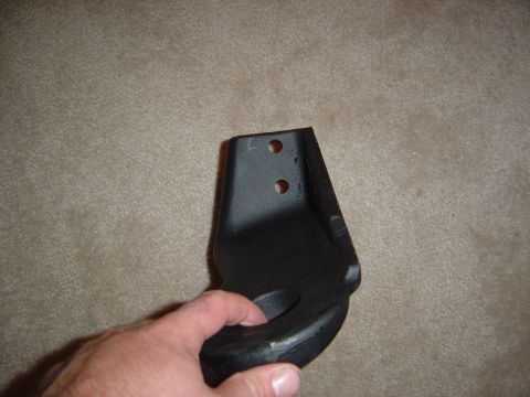 1986 Ford Bronco - A hand holds a black painted steel bracket with two mounting holes at the top and a curved notch at the bottom, photographed over beige car…