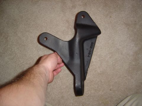 1986 Ford Bronco - A hand holds up a black metal bracket with two bolt holes and raised cast markings reading '1-04-1000', photographed against beige carpet.…