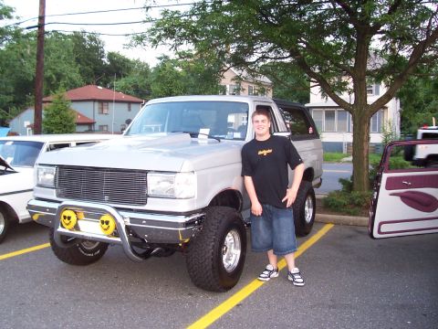 1990 Ford Bronco - A lifted Ford Bronco is parked in a lot with other vehicles and a young man standing beside it.