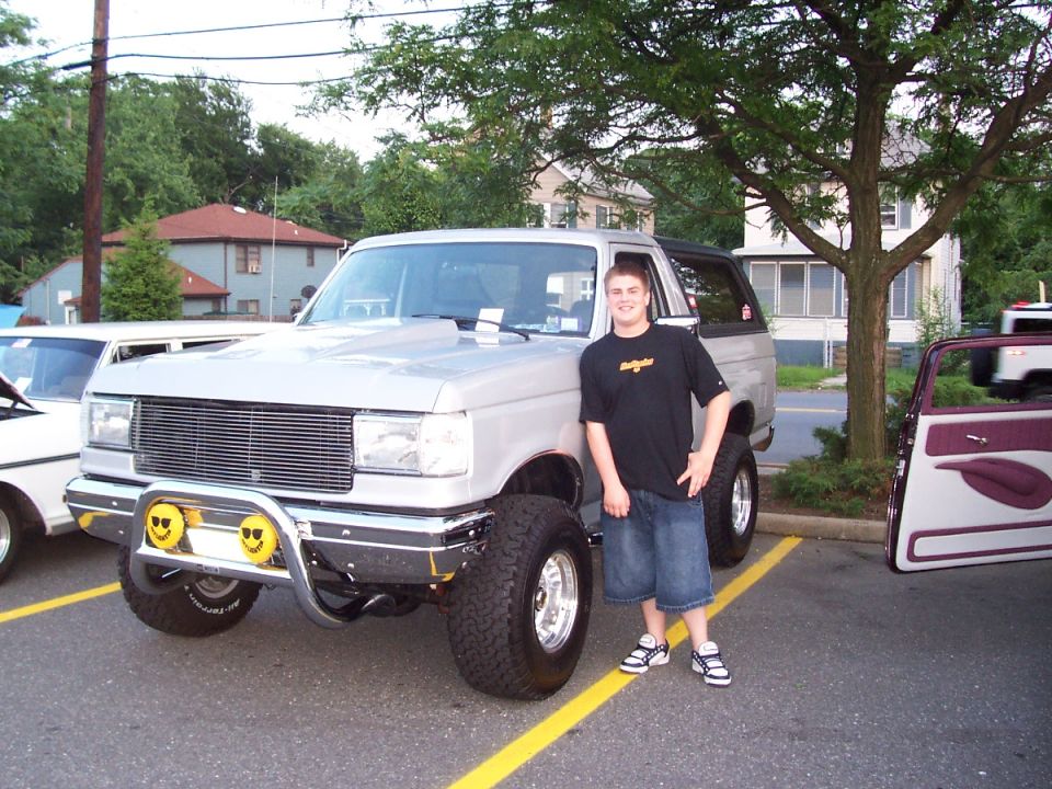 Silver lifted Ford Bronco with custom grille and smiley face fog lights parked next to a white car and a car with open doors