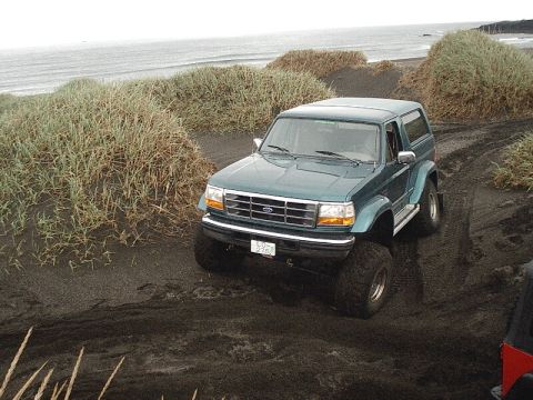 1996 Ford Bronco - This Ford Explorer is navigating a sandy terrain with coastal vegetation and ocean waves in the background.