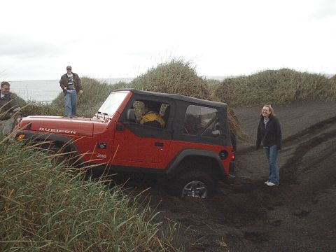 1996 Ford Bronco - A red Jeep Wrangler Rubicon is partially submerged in sand, with several people observing the scene near the coast.