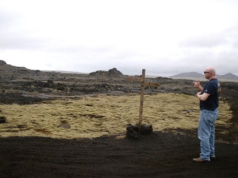 1996 Ford Bronco - A man gestures in a vast, rocky terrain under an overcast sky, with a simple wooden cross marking the spot.