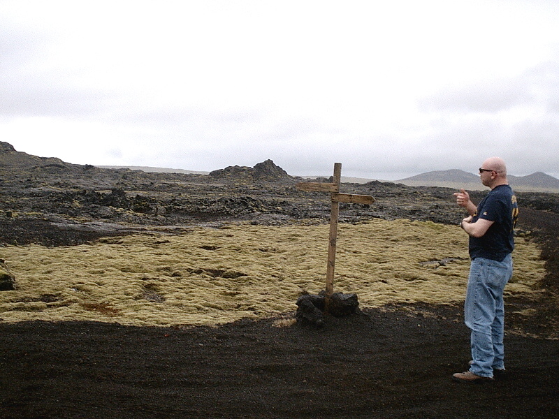 Man in sunglasses standing by a wooden cross in a barren volcanic field