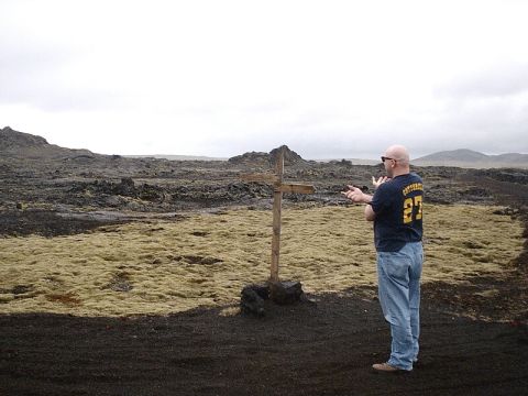 1996 Ford Bronco - A man stands near a wooden cross in a desolate, rocky area with sparse vegetation and a cloudy sky overhead.