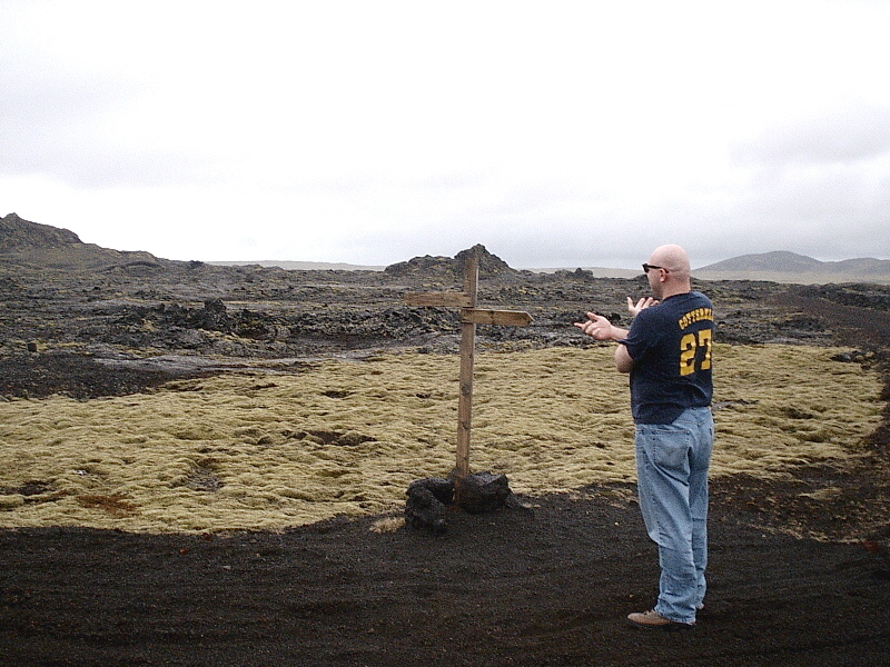 Man in sunglasses gesturing next to a wooden cross in a mossy volcanic field