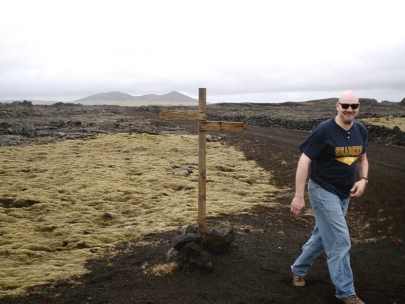 Man in sunglasses and SEABEES shirt walks past wooden signpost in a mossy volcanic landscape