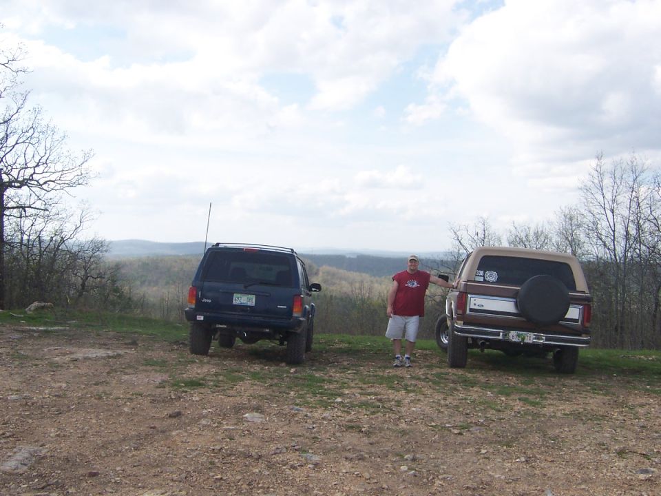 Rear of brown and tan Ford Bronco with spare tire cover parked beside blue Jeep Cherokee at a hilltop overlook
