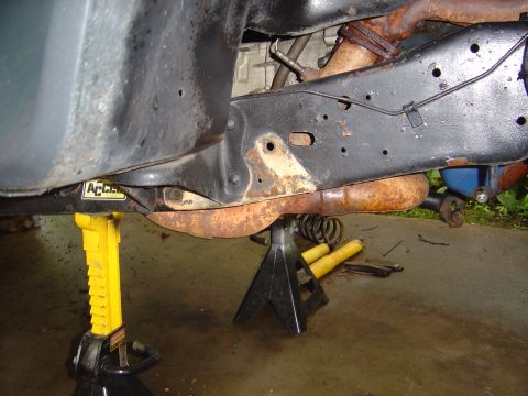 1986 Ford Bronco - Underside view of a vehicle's frame rail supported by a yellow ratchet jack stand and a black jack stand, showing a rusty exhaust pipe and…