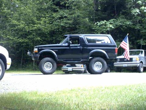 1995 Ford Bronco - A lifted black Ford Bronco is parked on a gravel surface next to a pickup truck flying an American flag.