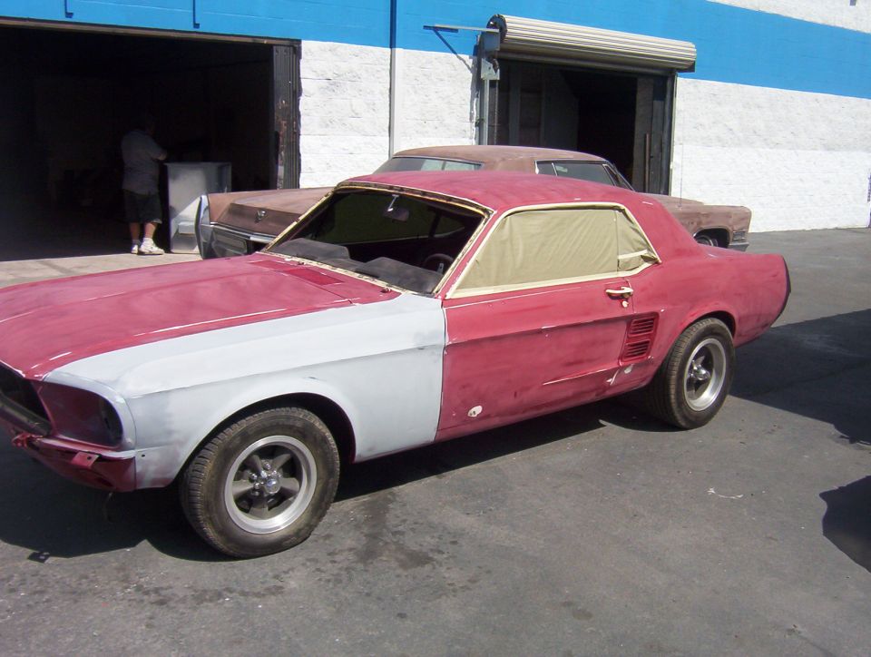 1967 Mustang coupe in red and gray primer with masked windows parked outside a body shop