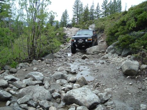 1989 Ford Bronco - A black 1989 Ford Bronco navigates a challenging, rocky off-road trail with another vehicle visible in the background.