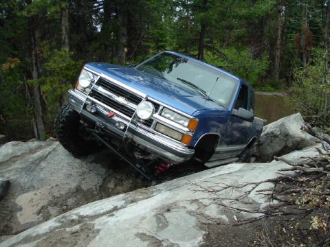 1989 Ford Bronco - Blue pickup truck is shown off-roading over large rocks on a wooded trail.
