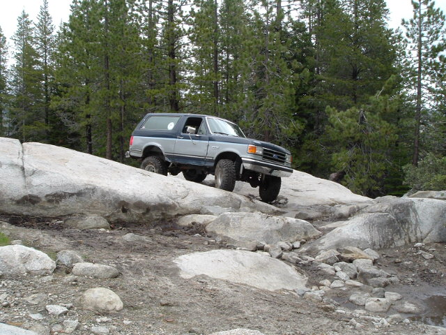 Blue and silver Ford Bronco descending a granite slab on a forest trail