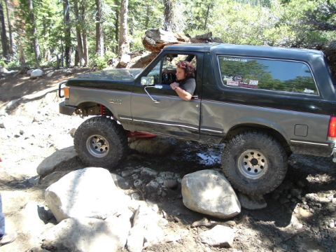 1989 Ford Bronco - A modified Ford Bronco tackles a challenging off-road course with large rocks and trees surrounding the path.