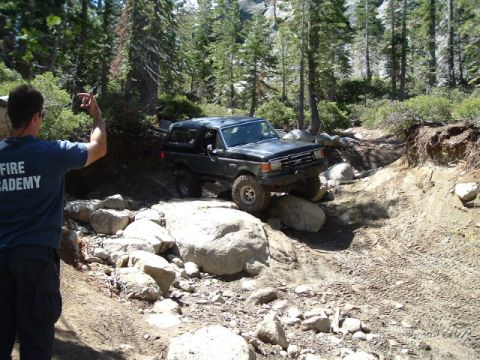 1989 Ford Bronco - A black Ford Bronco tackles a challenging rocky off-road course in a wooded area.