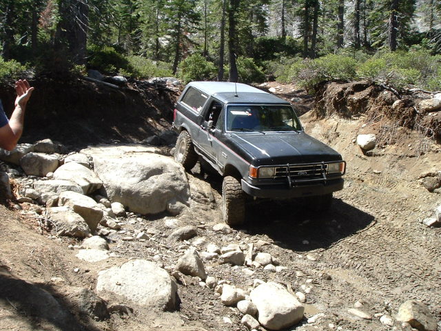 Blue and silver Ford Bronco stepping off a rock ledge on a dusty forest trail