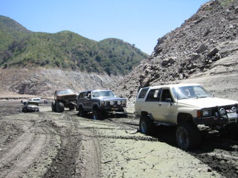 1985 Ford Bronco - Several off-road vehicles navigate a muddy terrain with steep, rocky hills in the background.