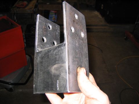 1978 Ford Bronco - A hand holds a pair of shop-fabricated bare-steel mounting brackets, each an angled plate with three drilled bolt holes and welded stiffeni…